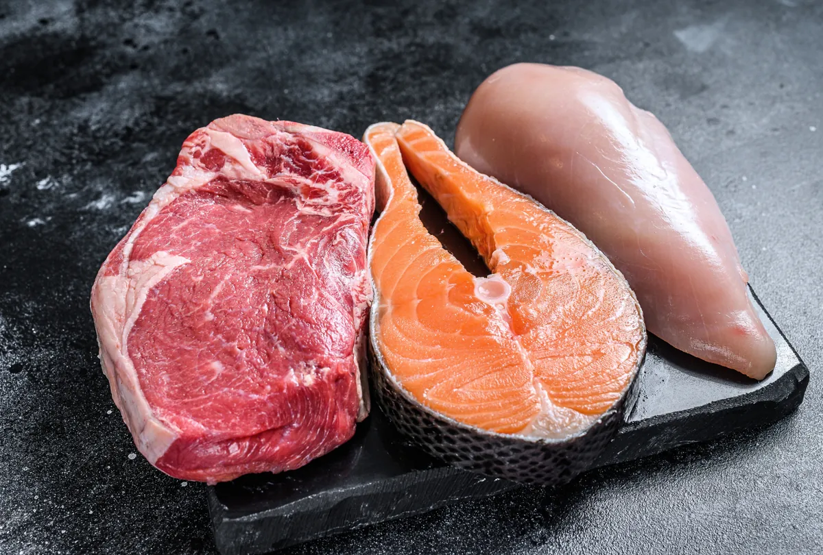 Raw beef steak, salmon fillet, and raw chicken breast on a black slate plate.